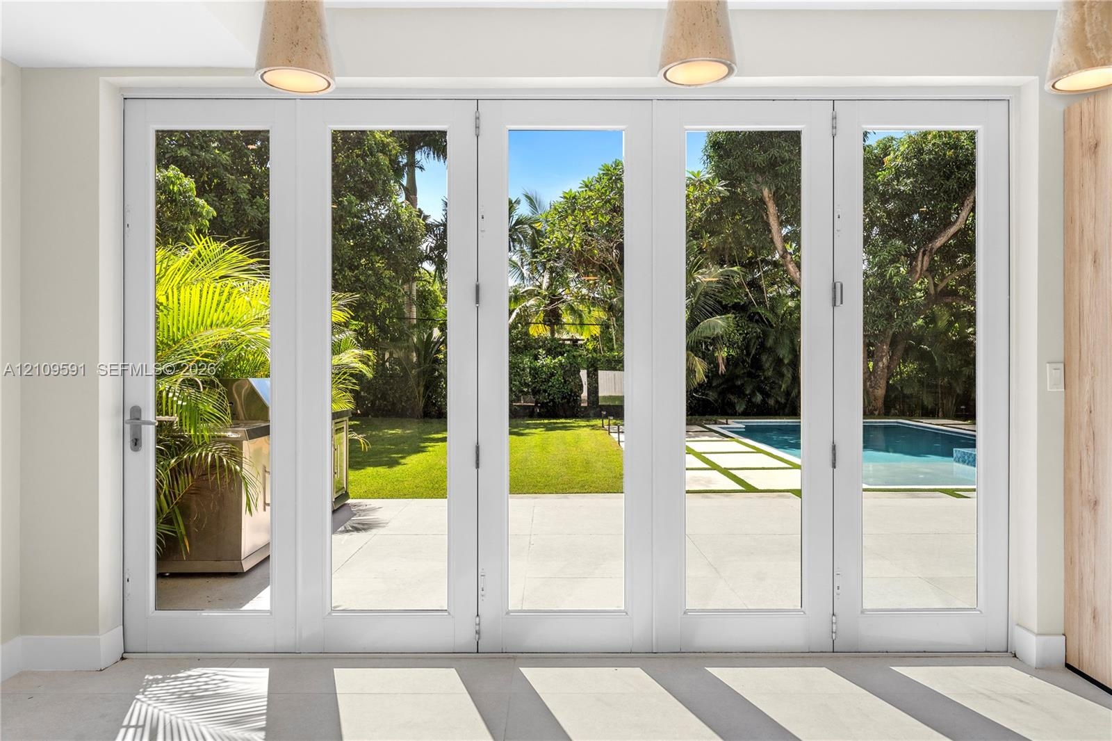 Multi-panel white-frame glass doors, stone patio, swimming pool, manicured lawn, and suspended pendant lighting