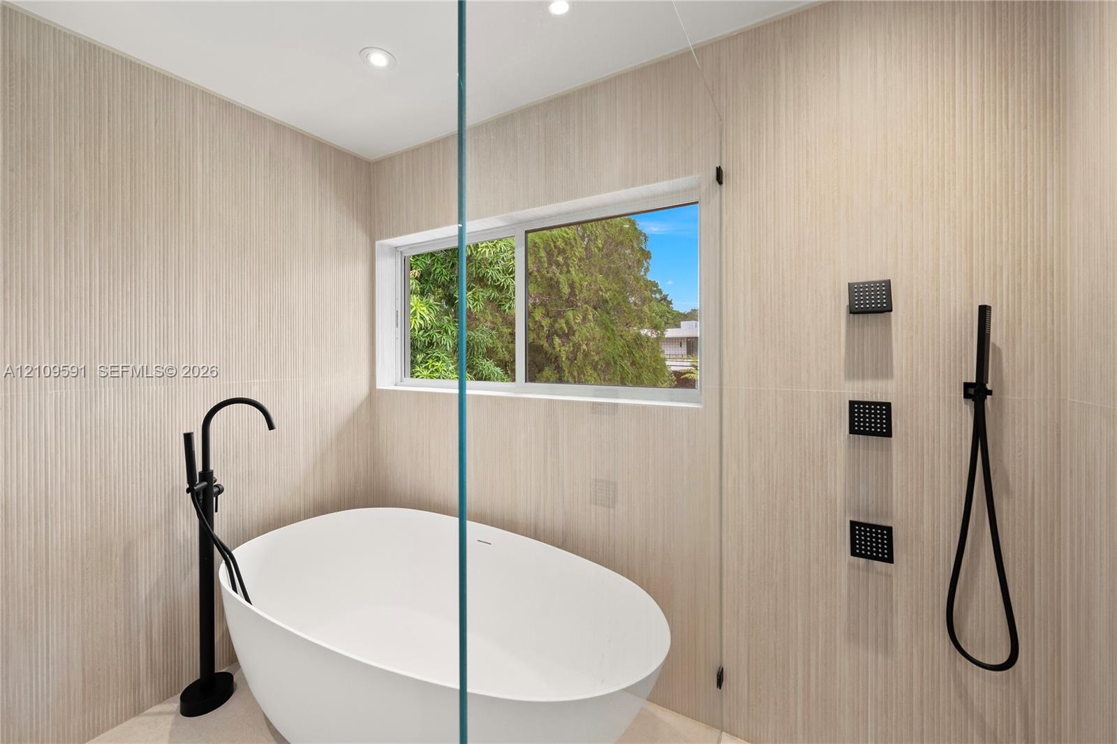 Freestanding white soaking tub with matte black floor-mounted faucet, floor-to-ceiling textured tile wall, frameless glass shower partition, and matte