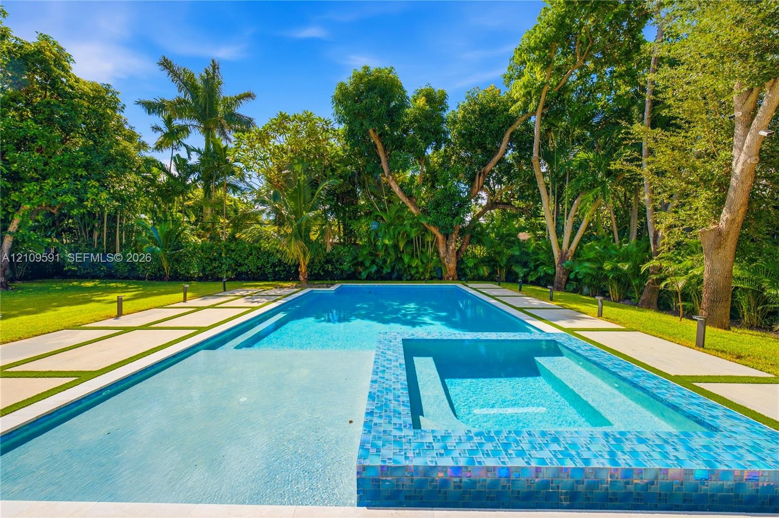In-ground swimming pool with an integrated spa, mosaic tile finish, concrete paver decking, and mature landscaping