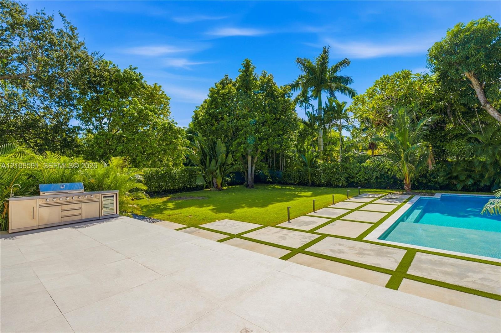 Backyard featuring a swimming pool, integrated stainless steel outdoor kitchen, stone paver patio with turf inlays, and mature landscaping