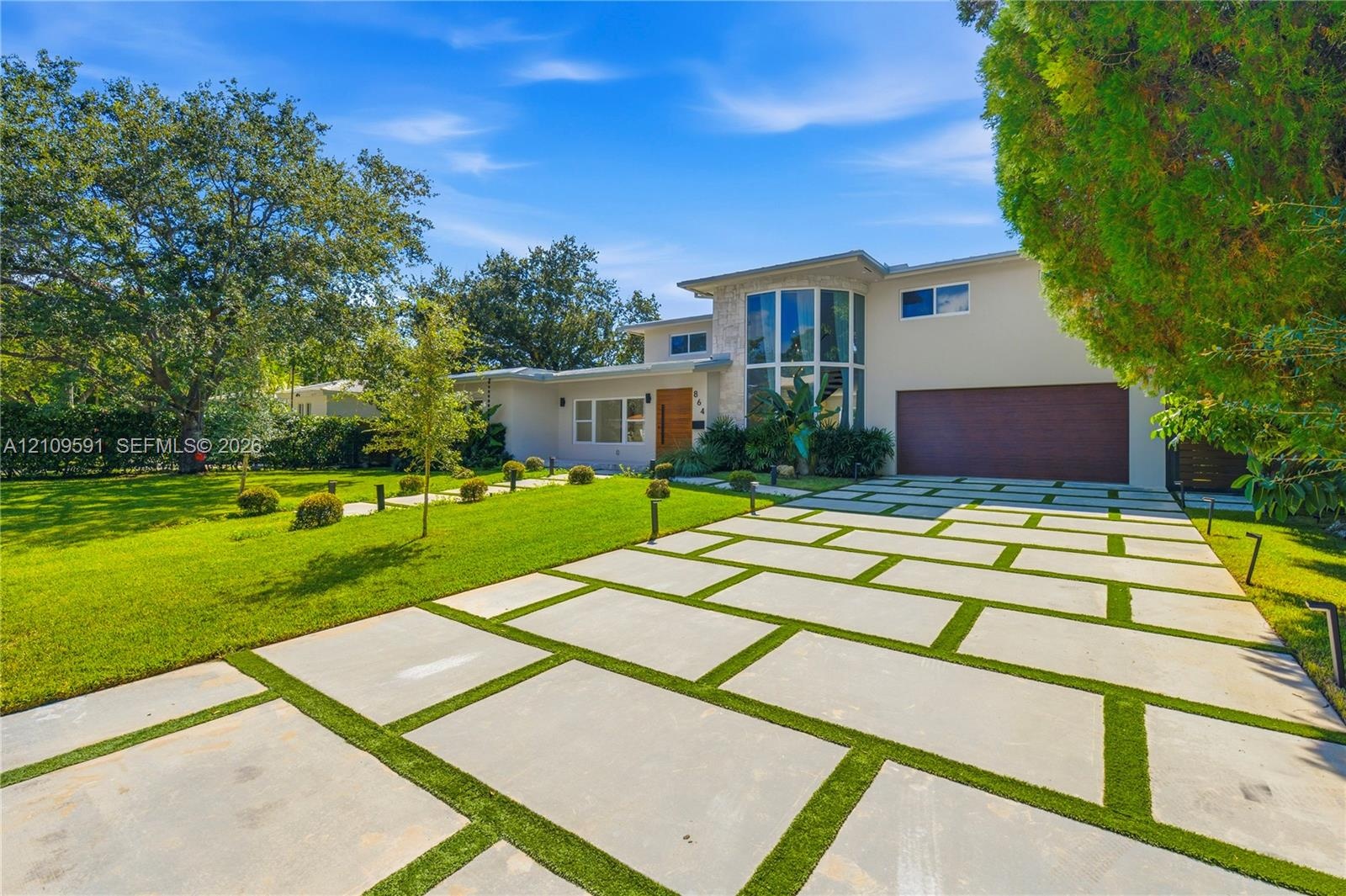 Contemporary residence featuring a segmented concrete paver driveway, vertical multi-story glass paneling, wood-finished entrance door, and a manicure
