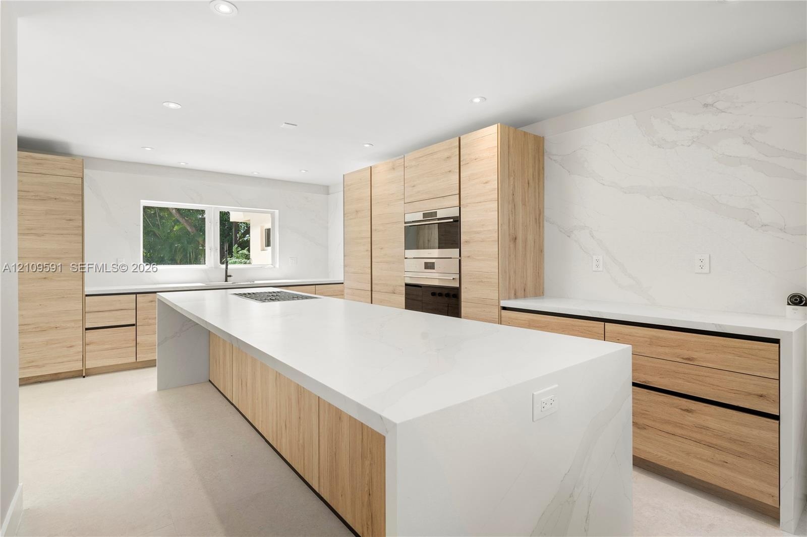 Modern kitchen featuring light wood-finish cabinetry, a waterfall-edge center island, solid-surface countertops, and full-height stone backsplash