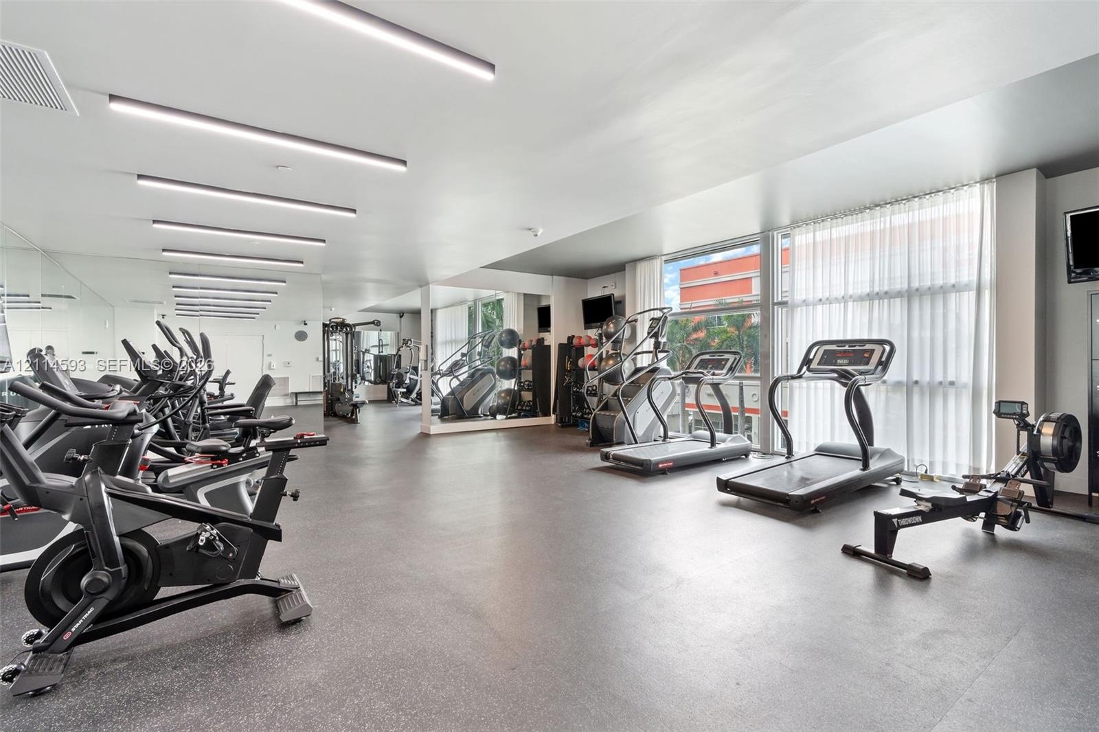 Fitness center featuring full-wall mirrors, recessed linear lighting, rubber flooring, and floor-to-ceiling windows with sheer drapery