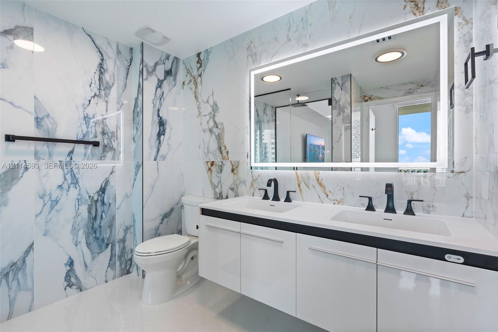 Floating white cabinetry with dual undermount sinks, matte black widespread faucets, large-format stone-finish wall tile, and an integrated LED-backli