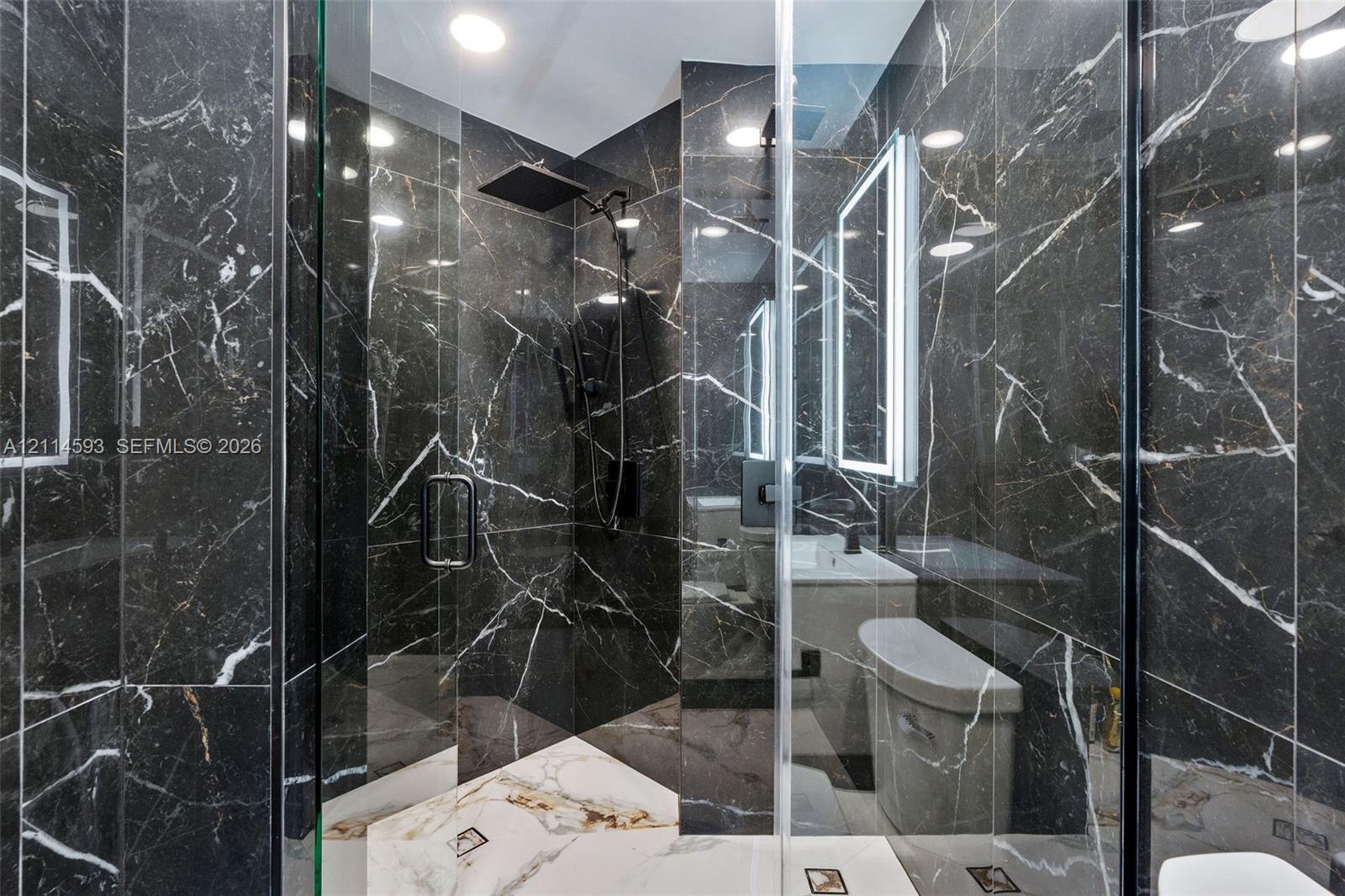 Walk-in shower featuring dark veined stone-finish tile surround, glass enclosure, square rainfall shower head, integrated LED-lit vanity mirror, and w