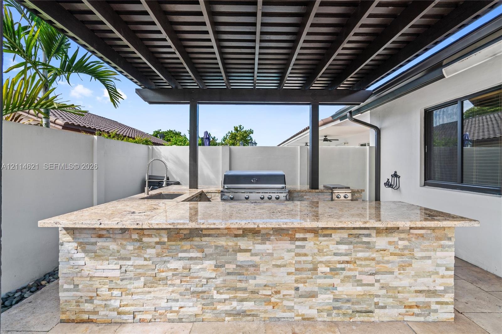 Outdoor kitchen featuring a stacked-stone base, natural stone countertop, built-in stainless steel grill, integrated sink, and a covered pergola struc