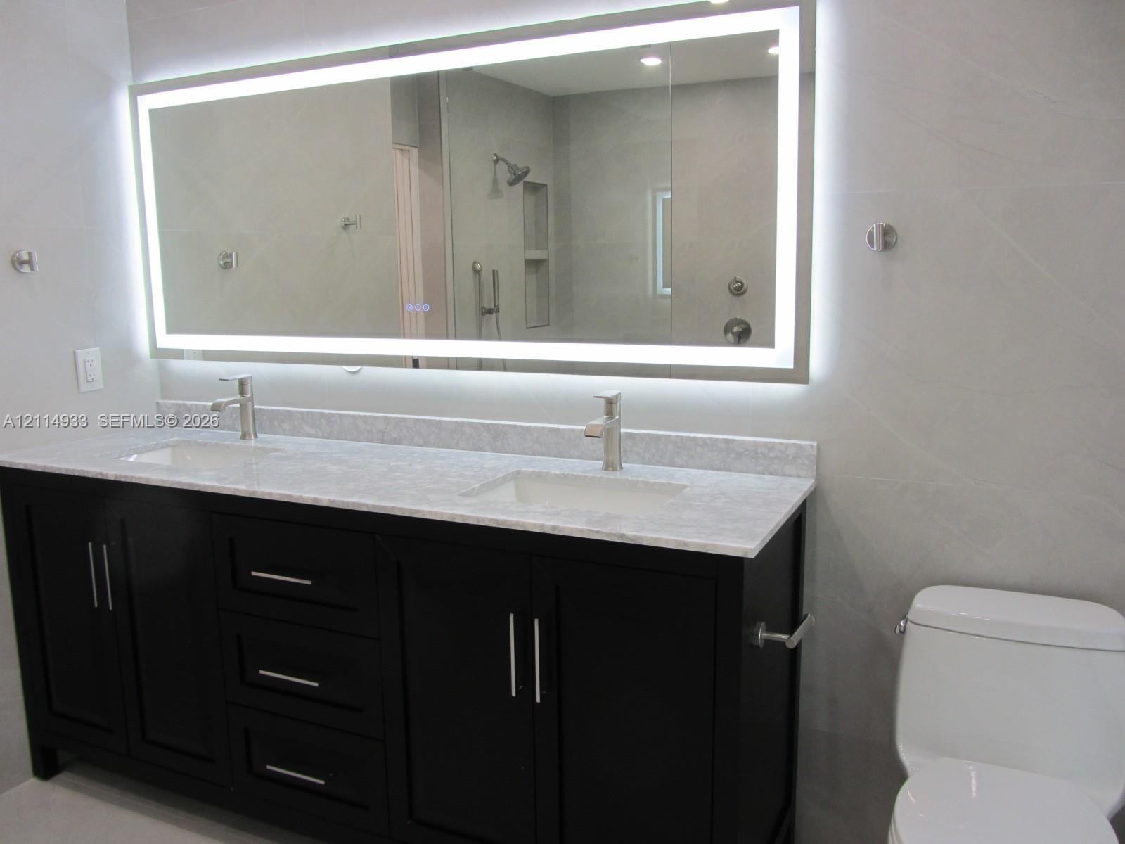 Dual-sink vanity featuring a stone countertop, oversized LED-backlit mirror, dark-finish cabinetry, and chrome-finish faucets