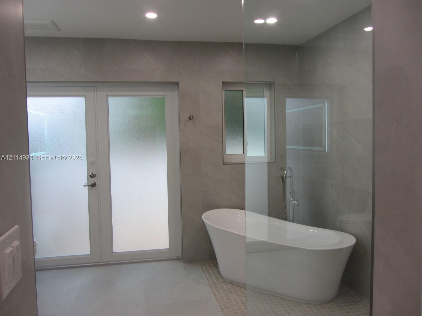 Freestanding soaking tub with a floor-mounted faucet, large-format grey tile wall cladding, glass shower enclosure, and white frosted glass double doo