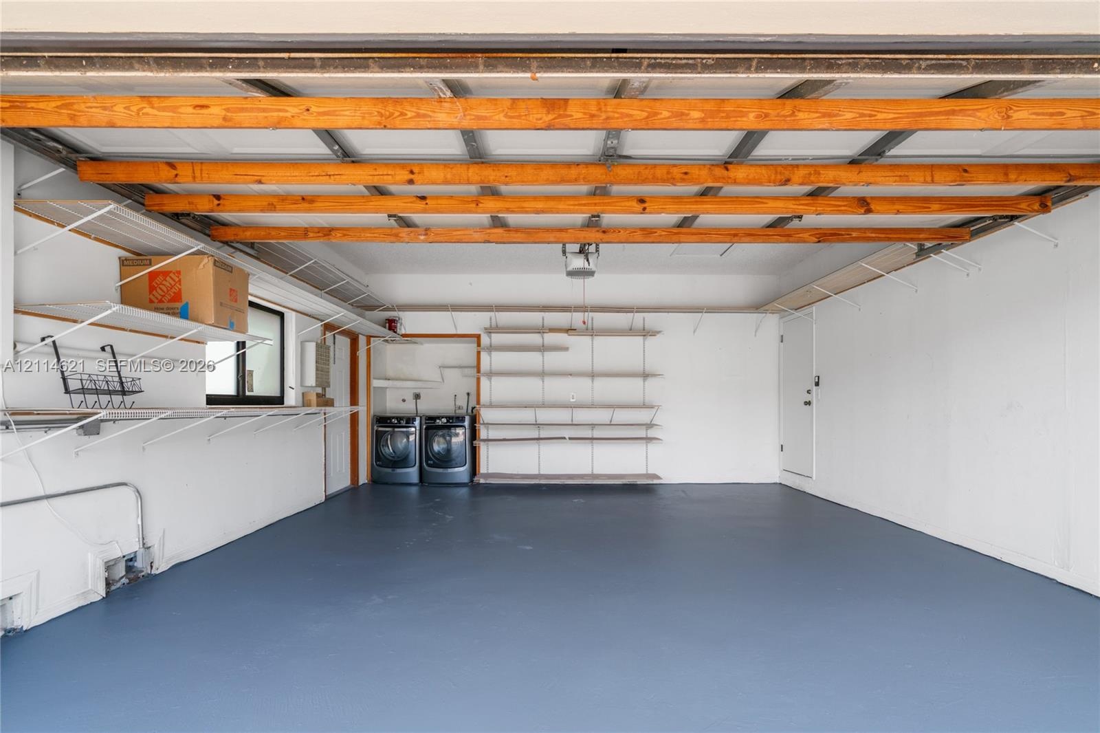 Garage interior featuring exposed wood ceiling beams, painted concrete flooring, mounted metal shelving units, and built-in laundry appliances