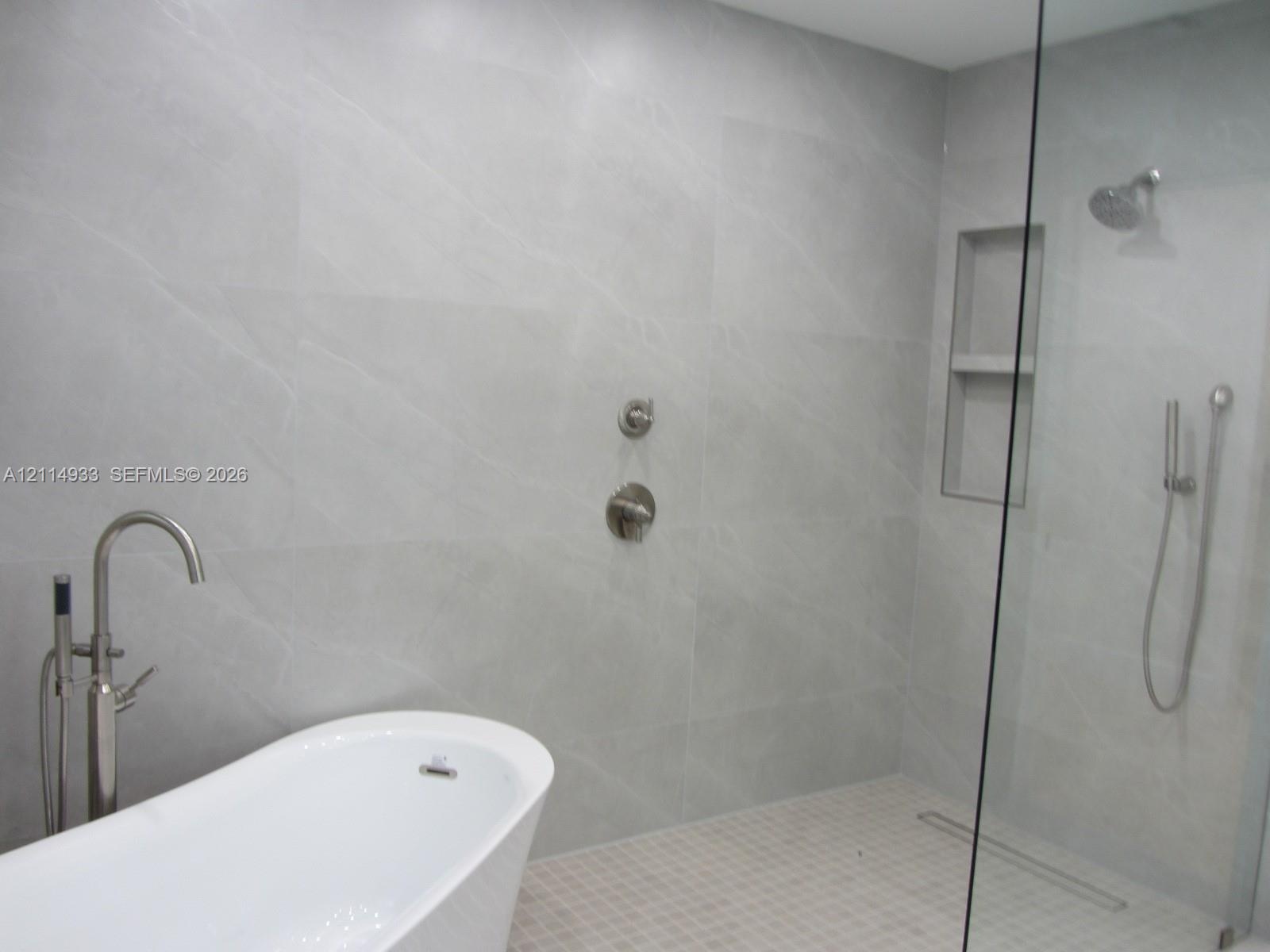 Freestanding soaking tub with floor-mounted faucet, walk-in shower featuring large-format tile surround, integrated tiled wall niche, rain shower head