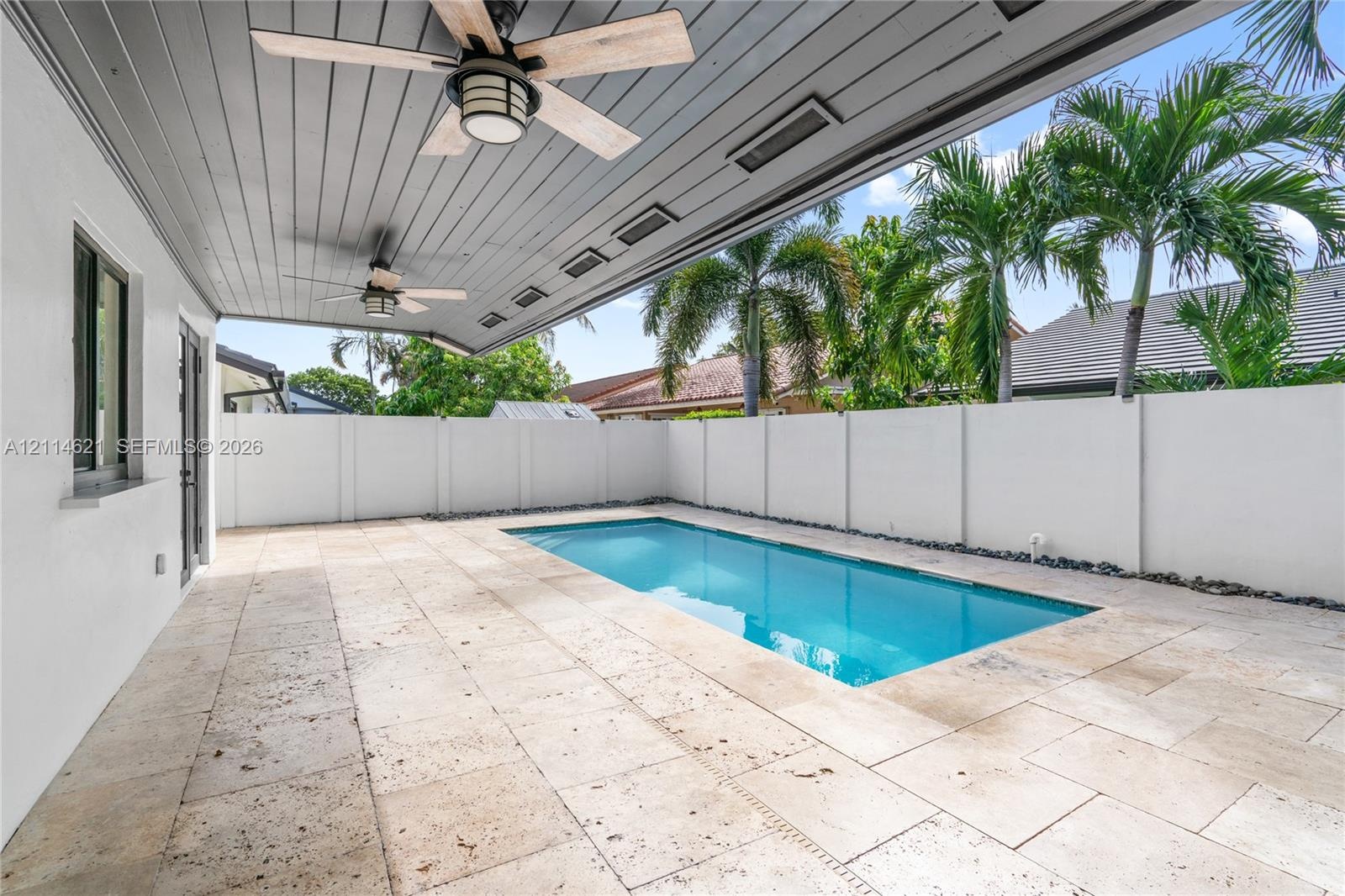 Covered patio featuring a grey tongue-and-groove ceiling with dual ceiling fans, stone paver decking, and a rectangular swimming pool enclosed by a wh