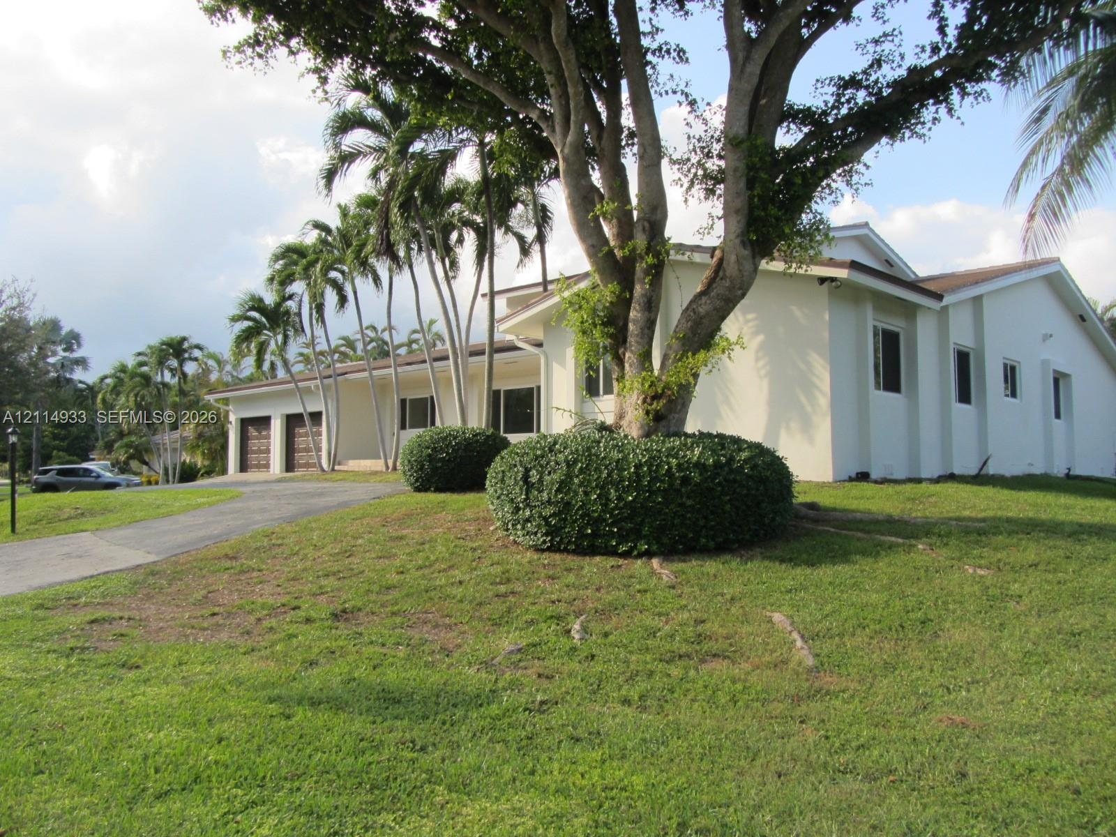 Stucco exterior featuring a two-car garage, expansive lawn, mature shade trees, and ornamental palms