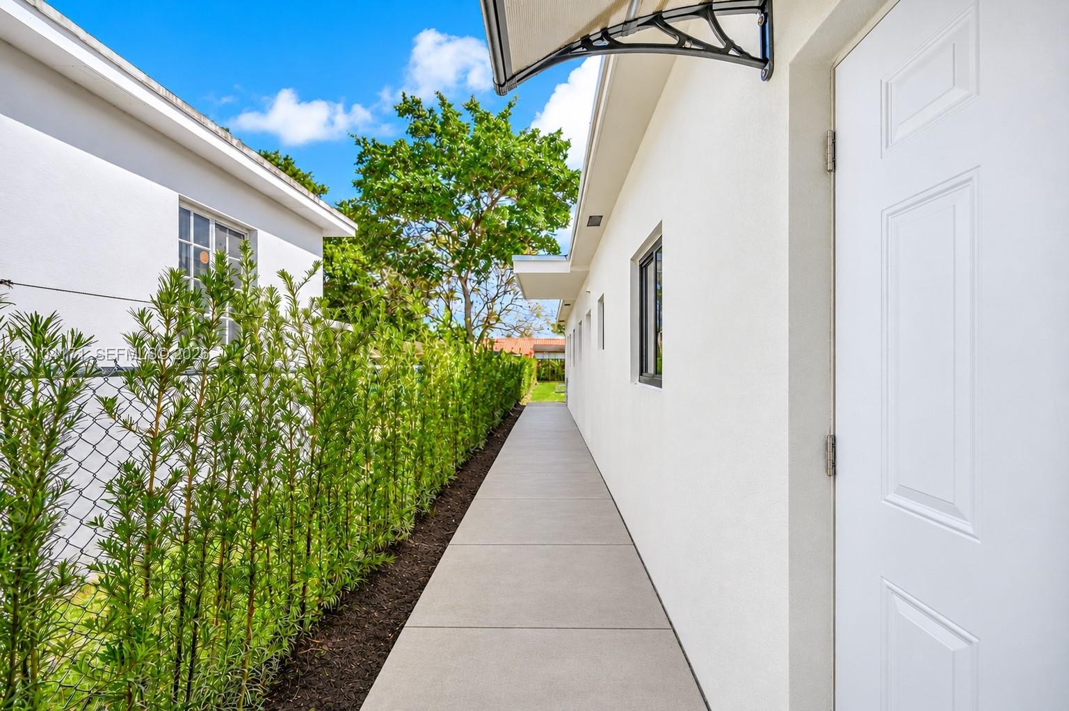 Exterior concrete walkway featuring a smooth stucco finish, a decorative metal door awning, and a manicured hedge border
