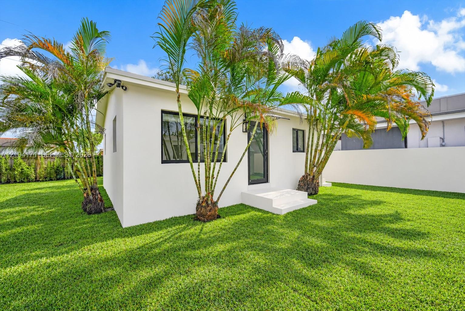 Stucco exterior featuring black-framed windows, a glass-panel entry door, concrete steps, and mature palm trees situated on a grassy lot enclosed by a