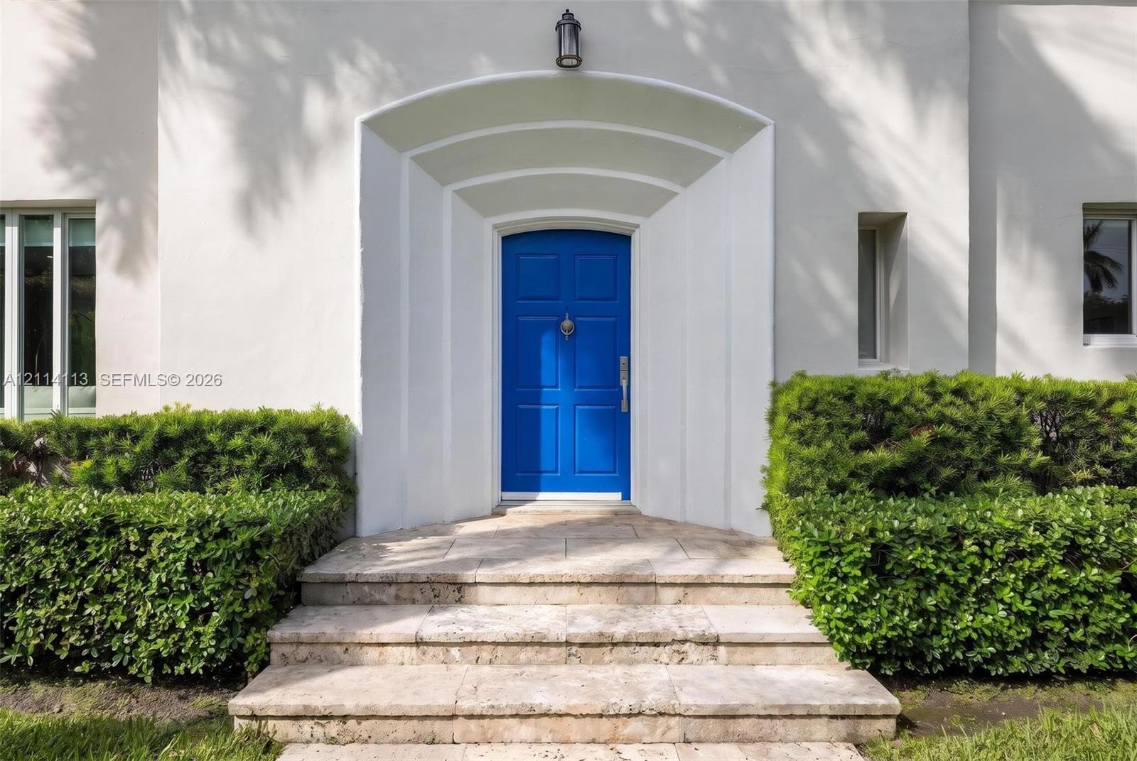 Stucco exterior entrance featuring an arched recessed doorway, paneled front door, stone steps, and manicured shrubbery