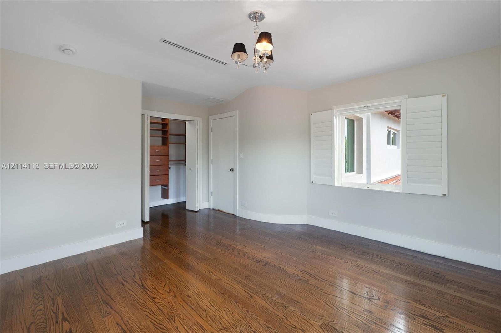 Room featuring wood-finish flooring, white plantation shutters, a multi-light chandelier, and a walk-in closet with built-in wooden shelving and drawe