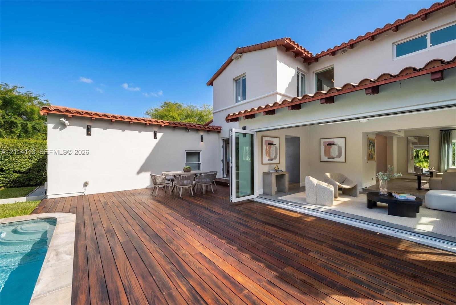Spanish-style exterior featuring a terra cotta tile roof, expansive wood deck, swimming pool with stone coping, and large-format bi-fold glass doors