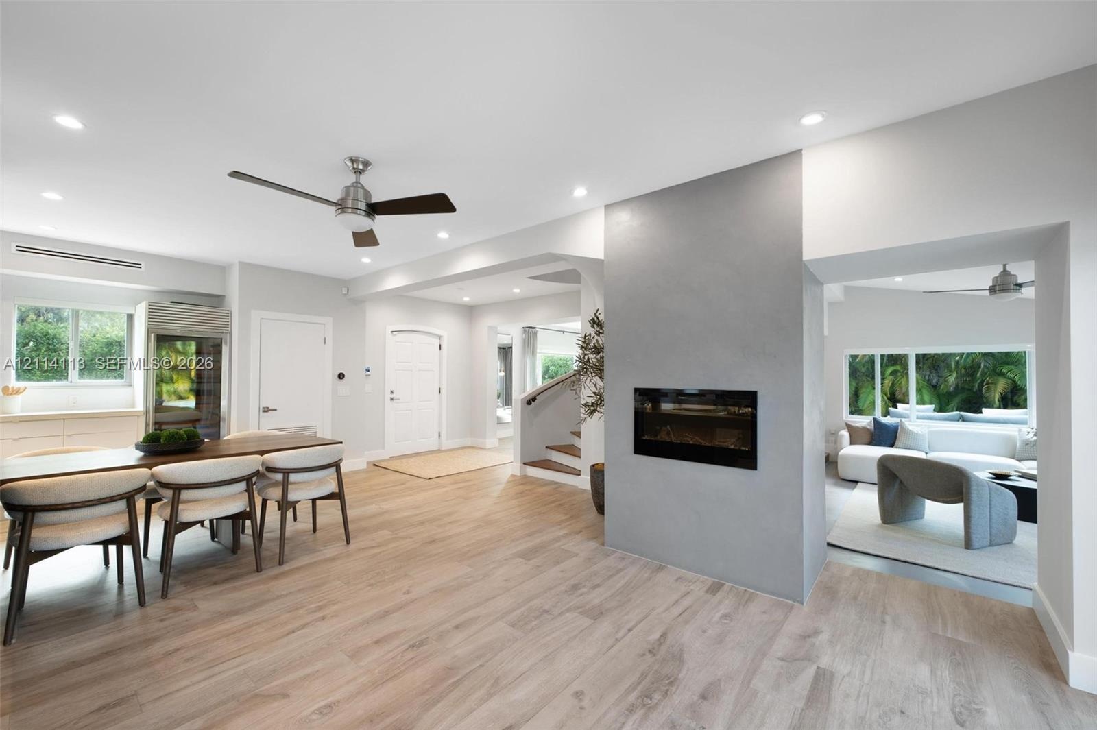 Light-toned wood-finish flooring, central fireplace with a grey smooth-finish surround, recessed ceiling lighting, ceiling fans, and multi-paned windo