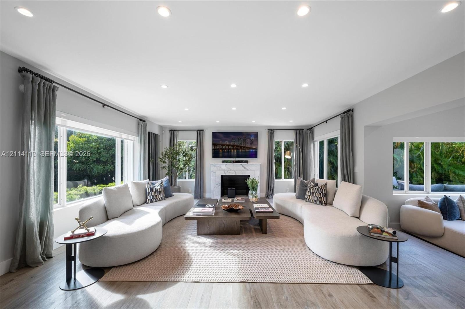 Living area featuring wood-finish flooring, recessed lighting, expansive multi-pane windows, and a central fireplace with a stone-finish surround