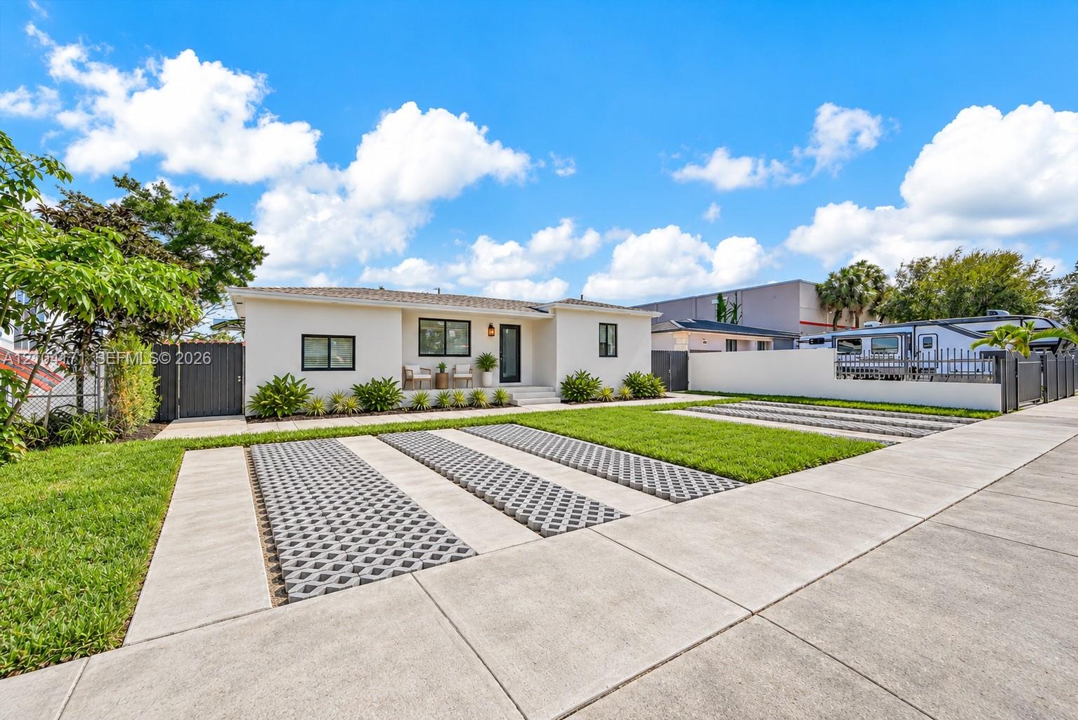 Single-story exterior with light-toned stucco finish, concrete driveway featuring permeable geometric pavers, manicured lawn, and opaque privacy fenci