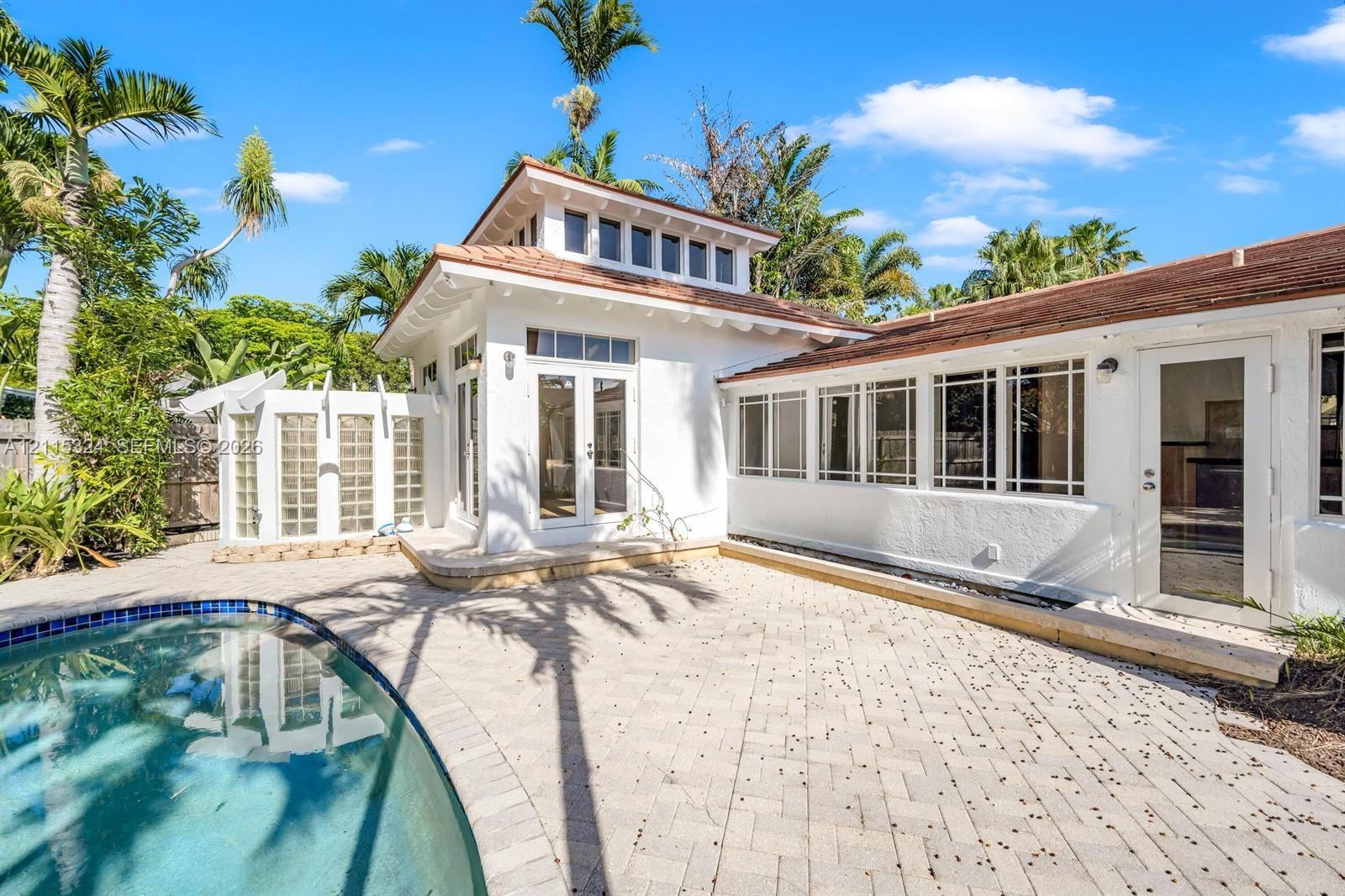 Exterior featuring white stucco siding, a curved in-ground pool, expansive stone-paver patio, glass-block privacy wall, and multi-pane transom windows