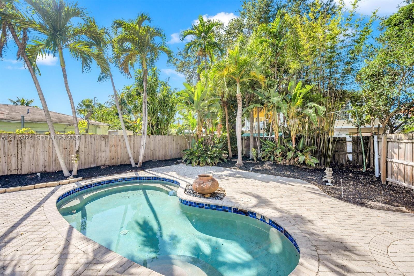 In-ground swimming pool with tile surround, expansive brick paver patio, and privacy wood fencing, framed by mature palms and lush landscaping