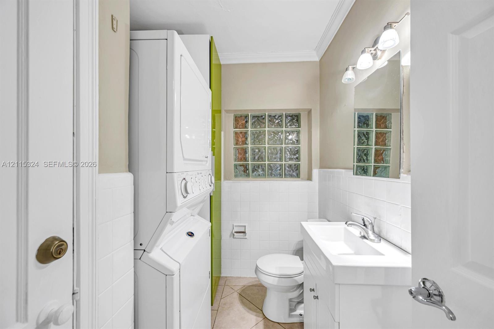 Combination laundry area and bathroom featuring a stacked washer and dryer, square glass block window, white vanity with integrated sink, and polished