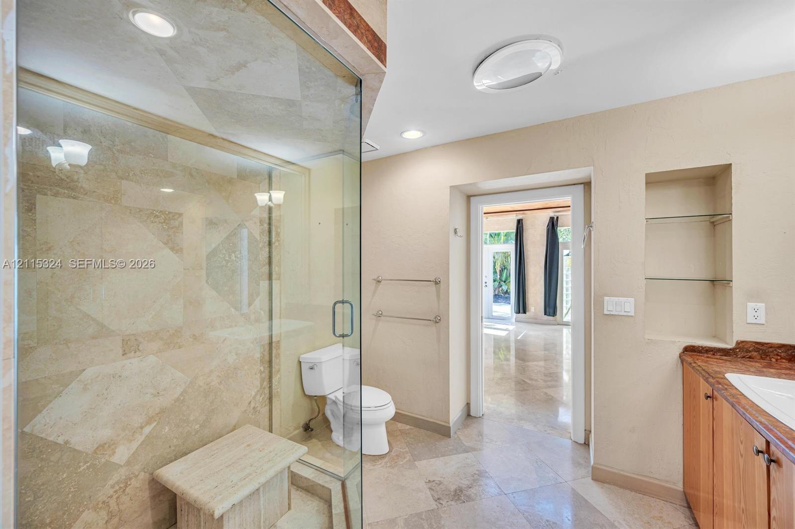 Walk-in shower with natural stone tile surround and built-in bench, glass enclosure, stone tile flooring, built-in wall shelving, and cabinetry with s