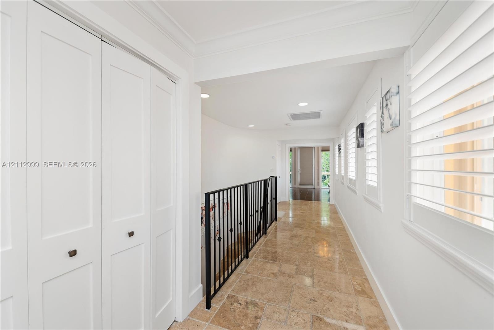 Hallway featuring stone tile flooring, white bi-fold closet doors, recessed lighting, plantation shutters, and a wrought-iron railing