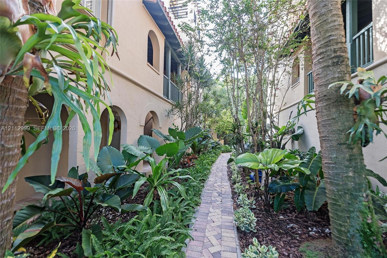 Stucco exterior featuring arched breezeways and second-story balcony railings, accented by a herringbone-patterned paver walkway and mature landscapin