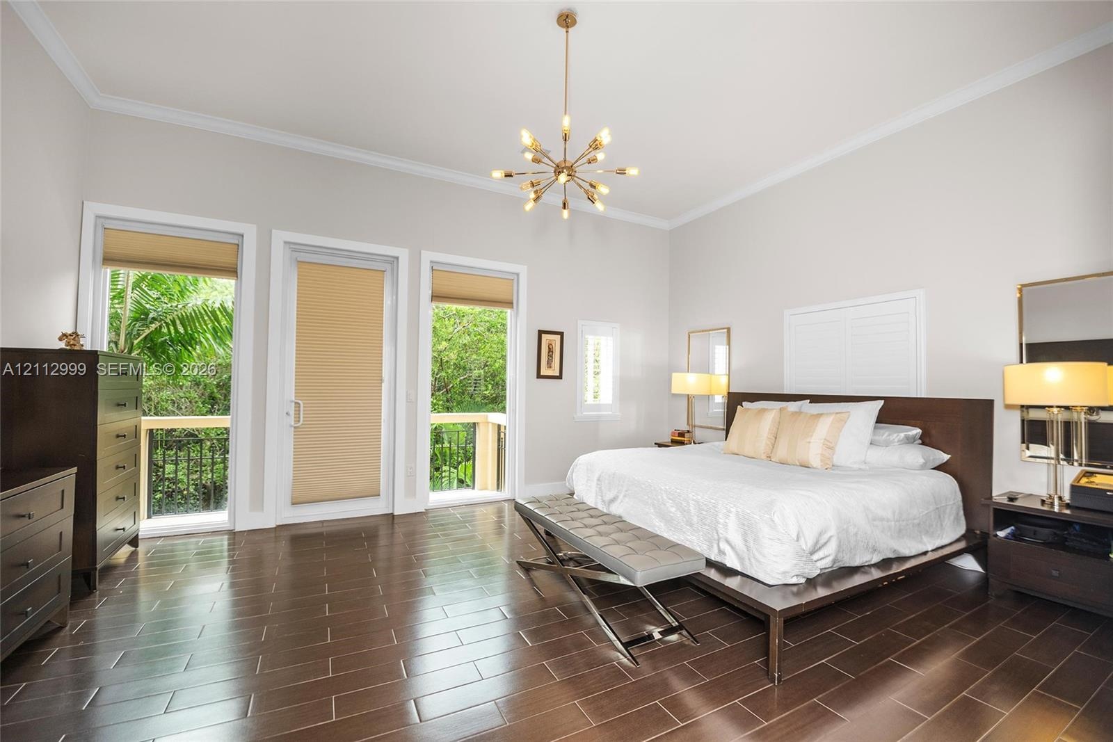 Spacious room featuring wood-finish tile flooring, crown molding, a central multi-arm chandelier, and two glass-panel doors leading to an exterior bal