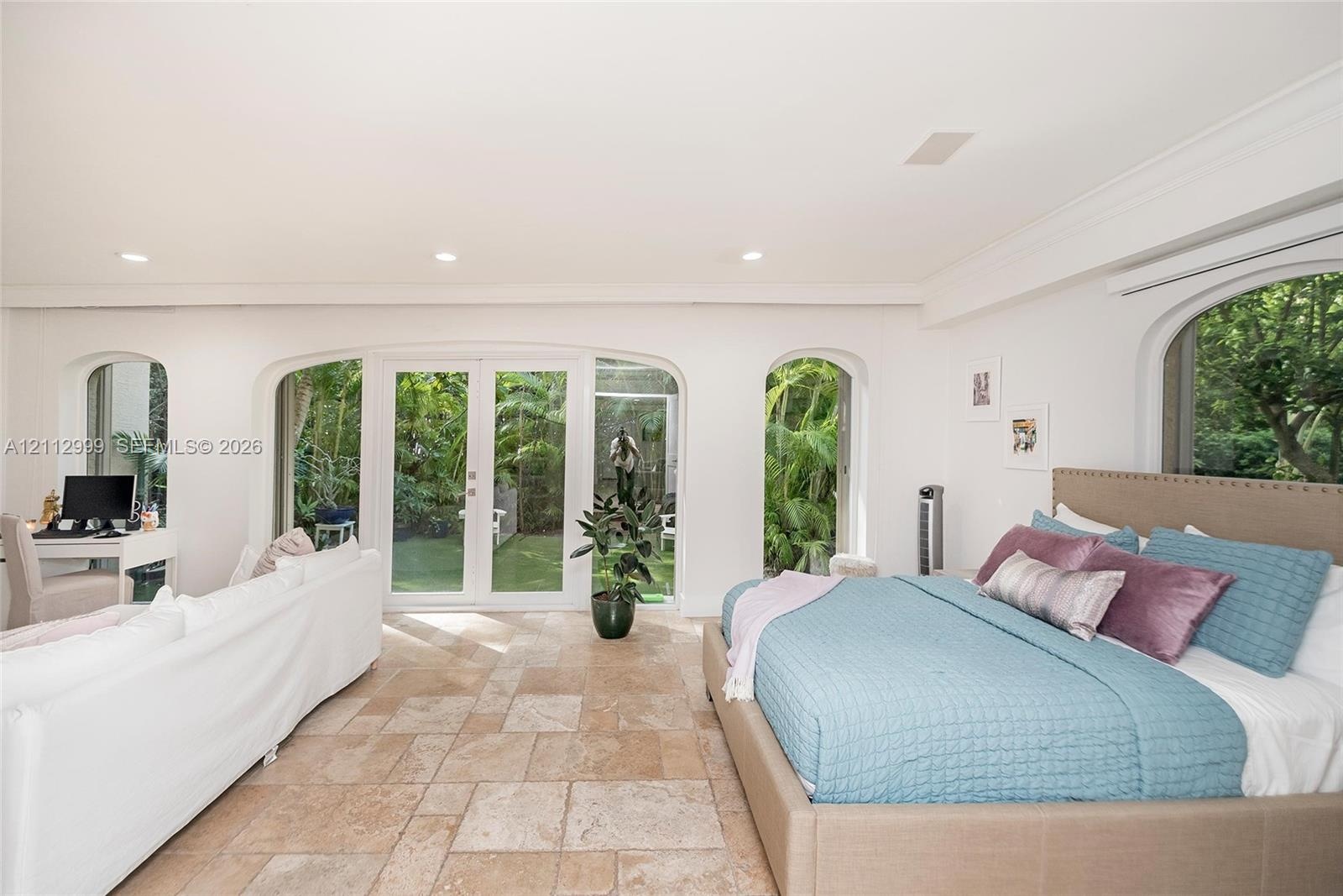 Spacious room featuring light stone-finish tile flooring, arched window casings, crown molding, and glass-paneled double doors providing outdoor acces