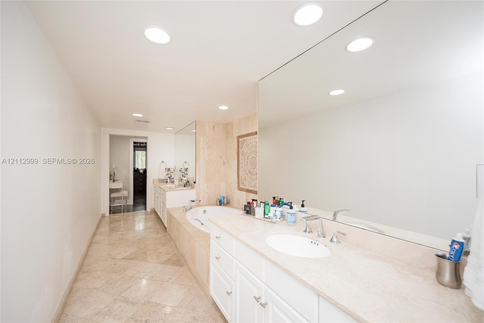 Bathroom featuring stone-finish flooring, a jetted soaking tub with a tiled surround, a lengthy stone-surface vanity with dual integrated sinks, and e