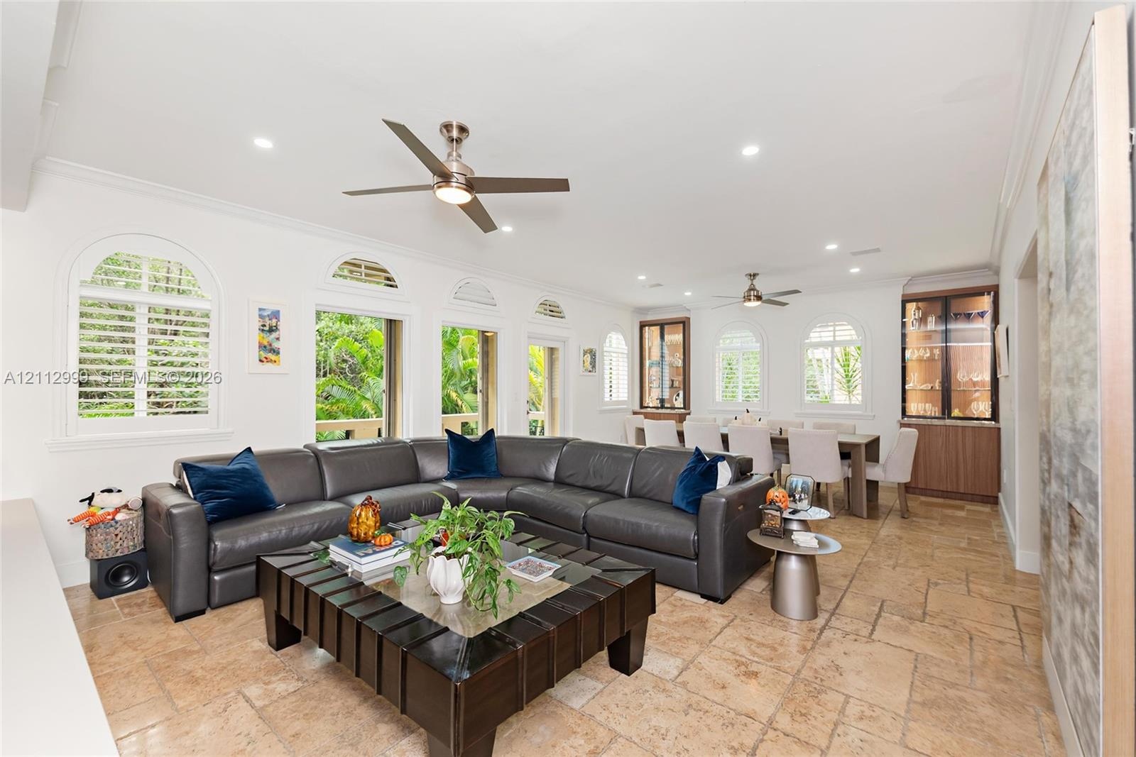 Open-concept interior featuring large-format tile flooring, multiple arched windows with built-in shutters, dual ceiling fans, and integrated custom c