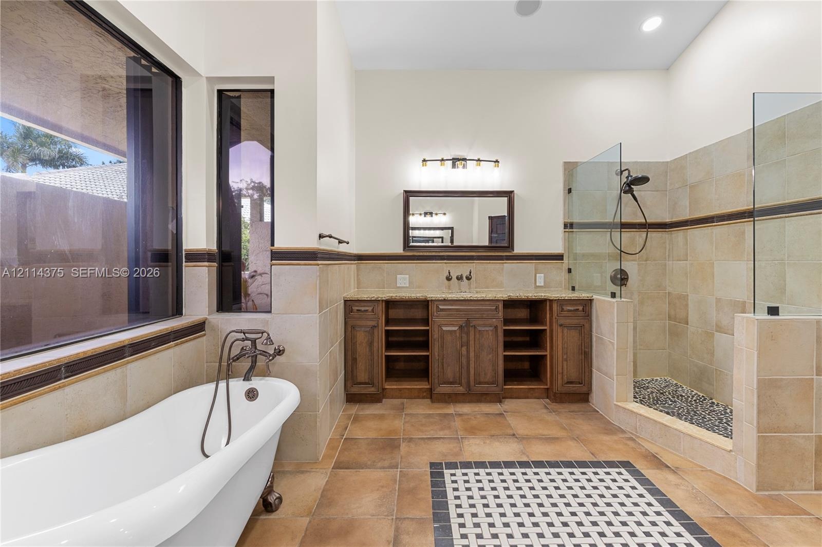 Freestanding soaking tub with dark metal faucet, dual-sink wooden vanity with stone countertop, tiled walk-in shower with glass enclosure, and pattern