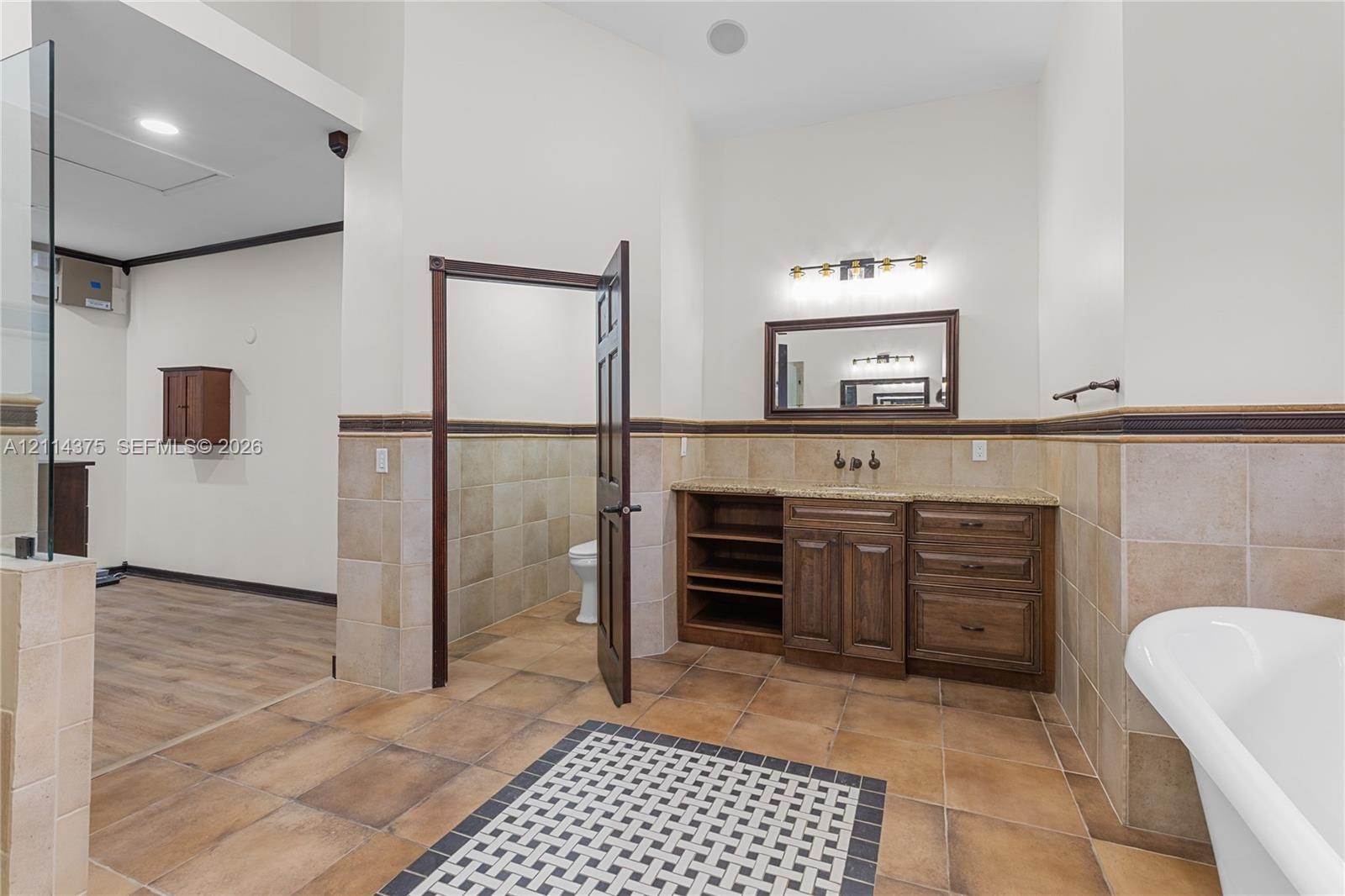 Bathroom featuring a wood-tone vanity with stone countertop, tile wainscoting, a wall-mounted framed mirror, a soaking tub, and a decorative tile inla
