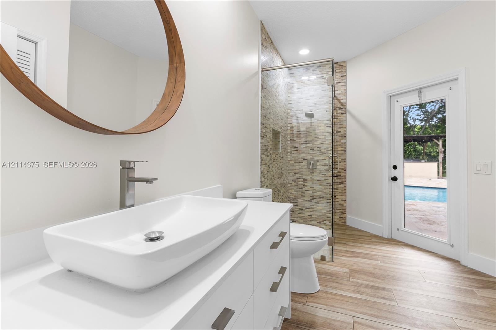 Bathroom featuring a rectangular vessel sink with a chrome faucet, wood-framed circular mirror, walk-in shower with stone-tile surround, wood-finish f
