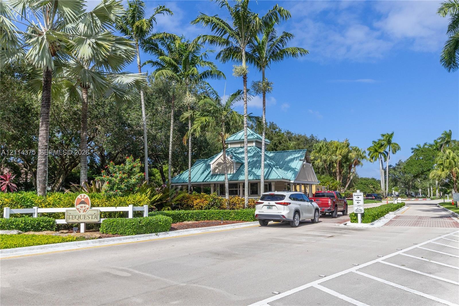 Gated community entrance featuring a teal metal roof structure, mature palm trees, white picket fencing, and manicured perimeter landscaping