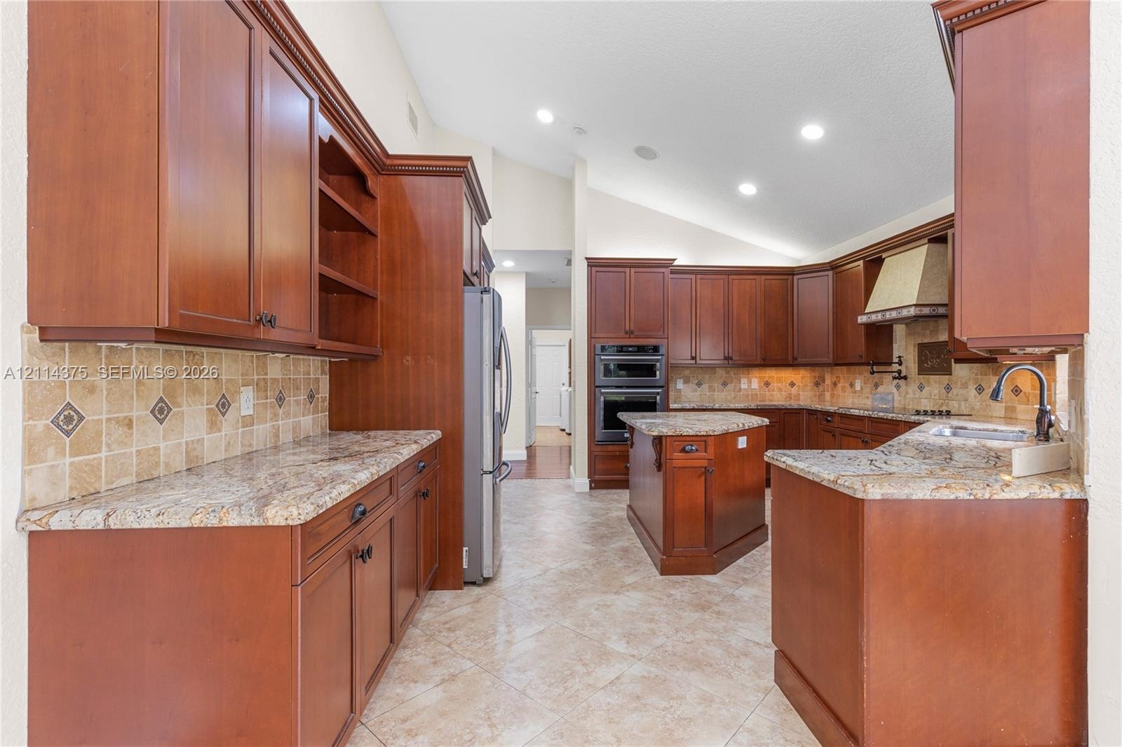 Kitchen featuring cherry-finish cabinetry, stone countertops, square-tile backsplash with decorative inserts, double wall oven, and center island