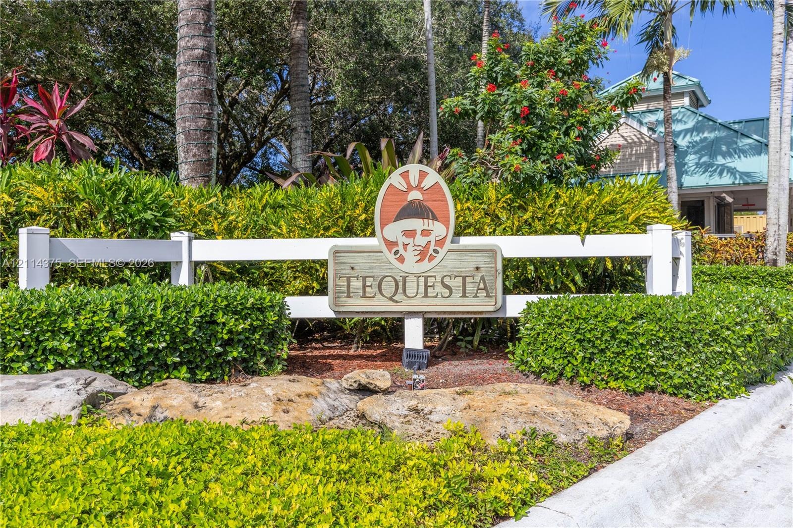 Community signage featuring custom wooden inlay, framed by a white post-and-rail fence, manicured hedges, and mature tropical landscaping