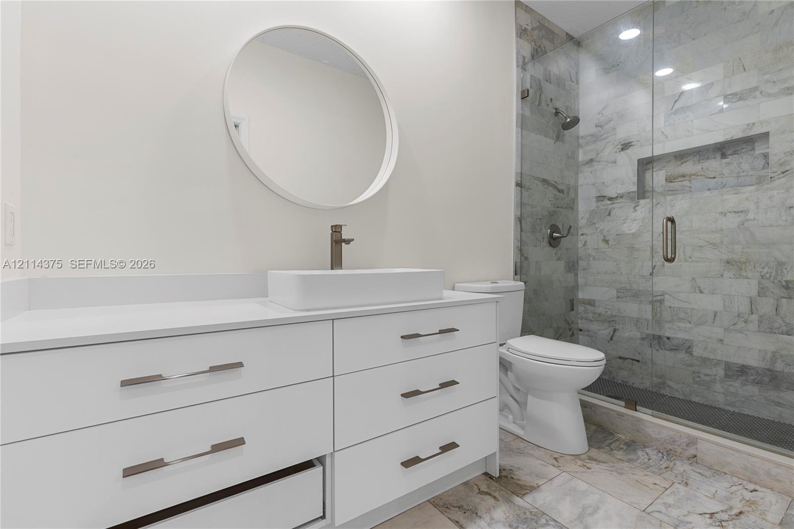 White cabinetry vanity with rectangular vessel sink, frameless glass shower enclosure with stone-tile surround, built-in shower niche, and round wall-