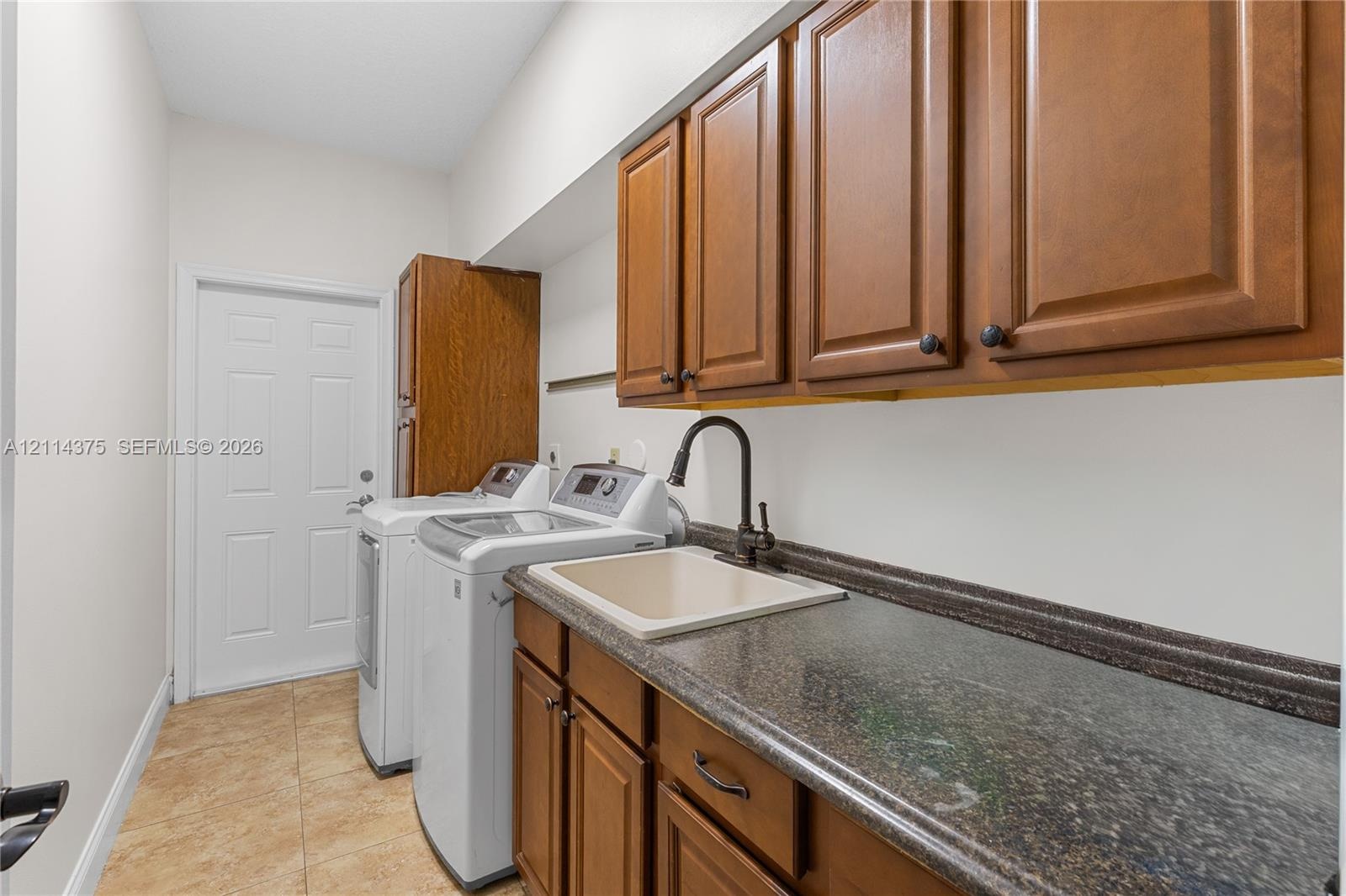 Laundry room featuring wood-finish cabinetry, stone-texture countertop, drop-in basin sink, matte black gooseneck faucet, and tile flooring