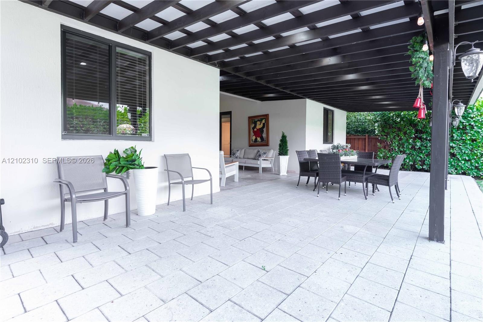 Covered patio featuring an expansive stone paver surface, dark-stained overhead pergola, wall-mounted exterior lanterns, and smooth stucco finish