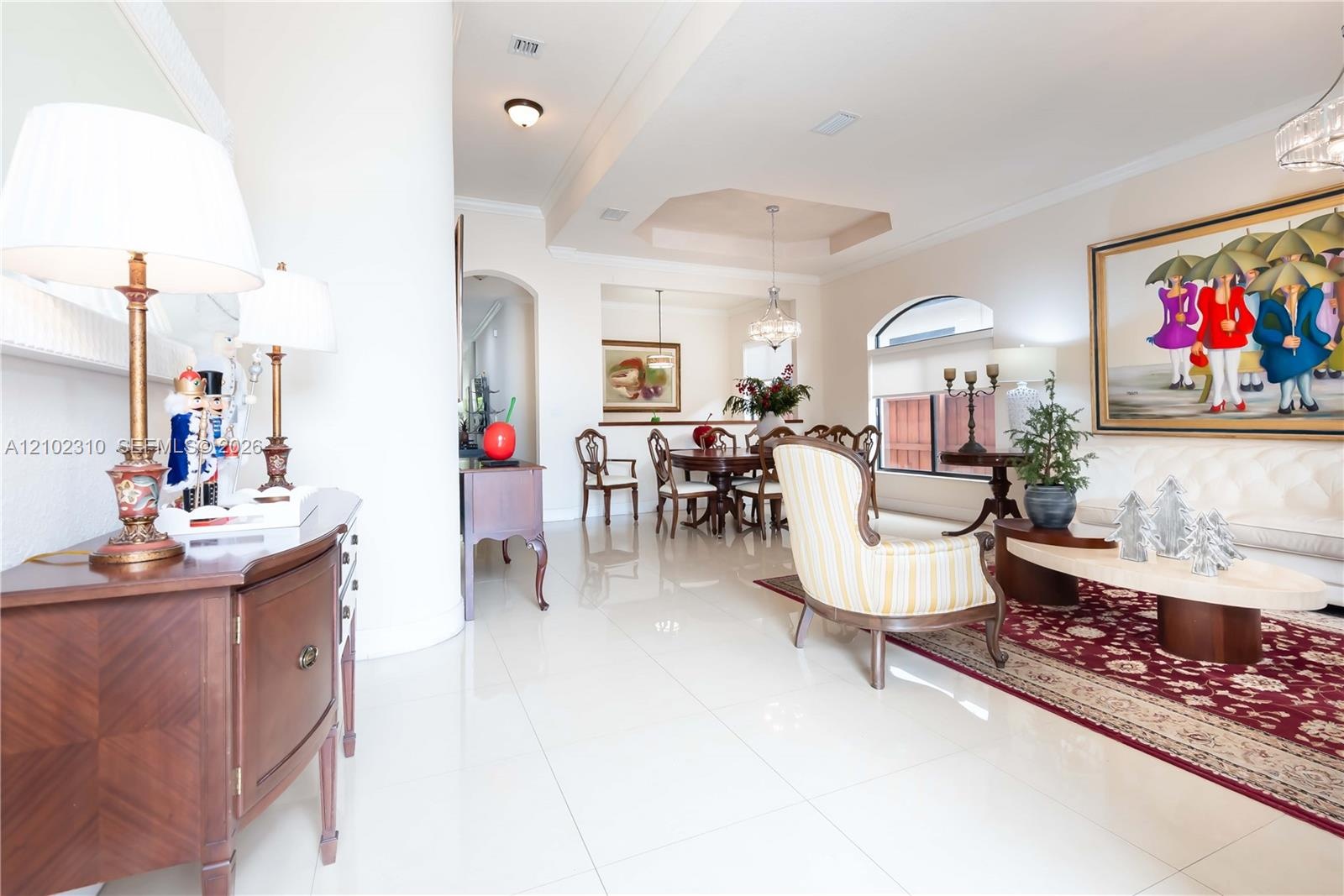 Open-concept interior featuring large-format tile flooring, recessed tray ceiling, arched architectural detailing, and ornate crystal chandelier light