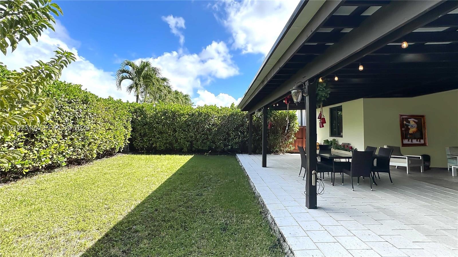 Covered patio featuring paver flooring, overhead support beams, recessed lighting, and a manicured lawn bordered by a tall privacy hedge