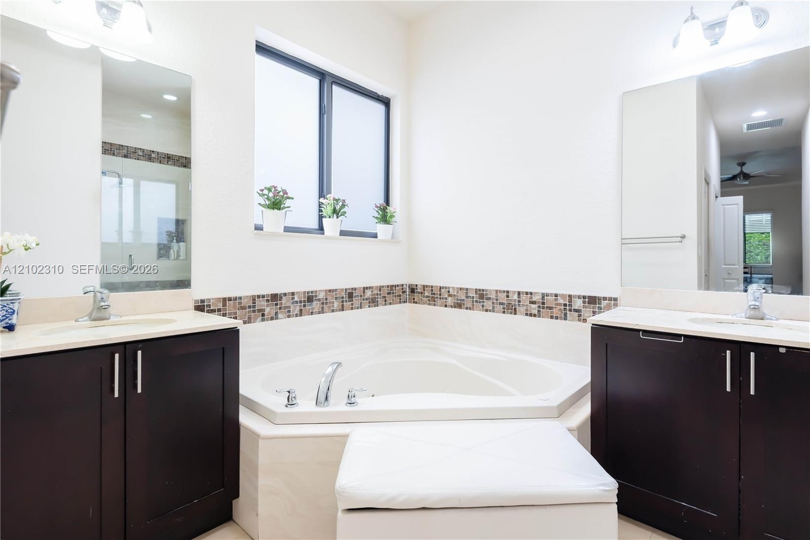 Dual dark wood cabinetry with neutral countertops, recessed corner soaking tub, tile backsplash, and oversized vanity mirrors
