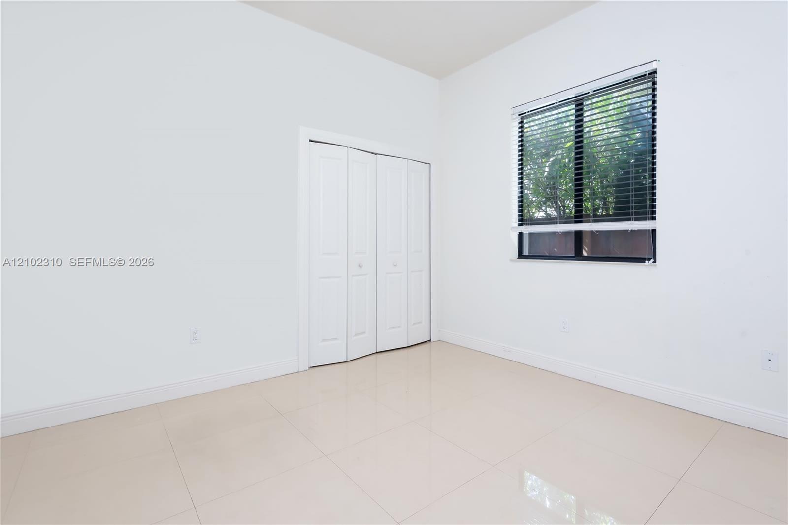 Neutral-toned tile flooring, white-panel bifold closet doors, and a single-pane window with horizontal blinds