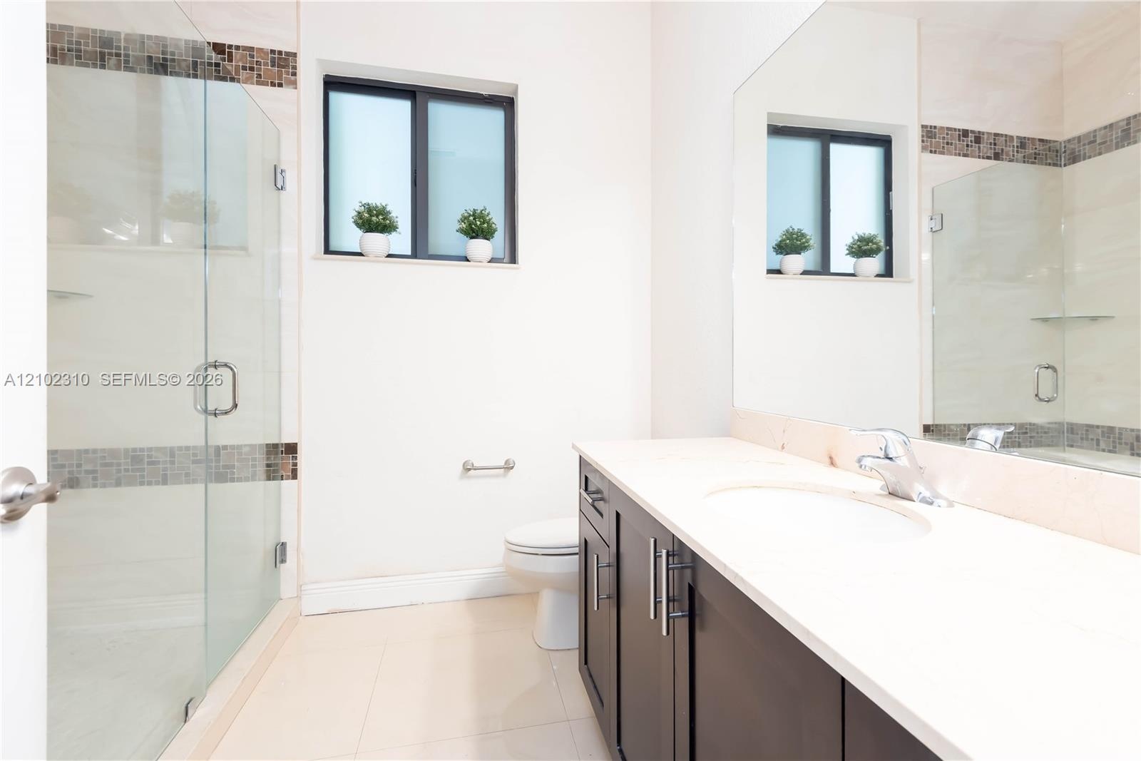 Walk-in shower with a glass enclosure, dark cabinetry with a solid-surface countertop, rectangular undermount sink, and mosaic tile wall accents