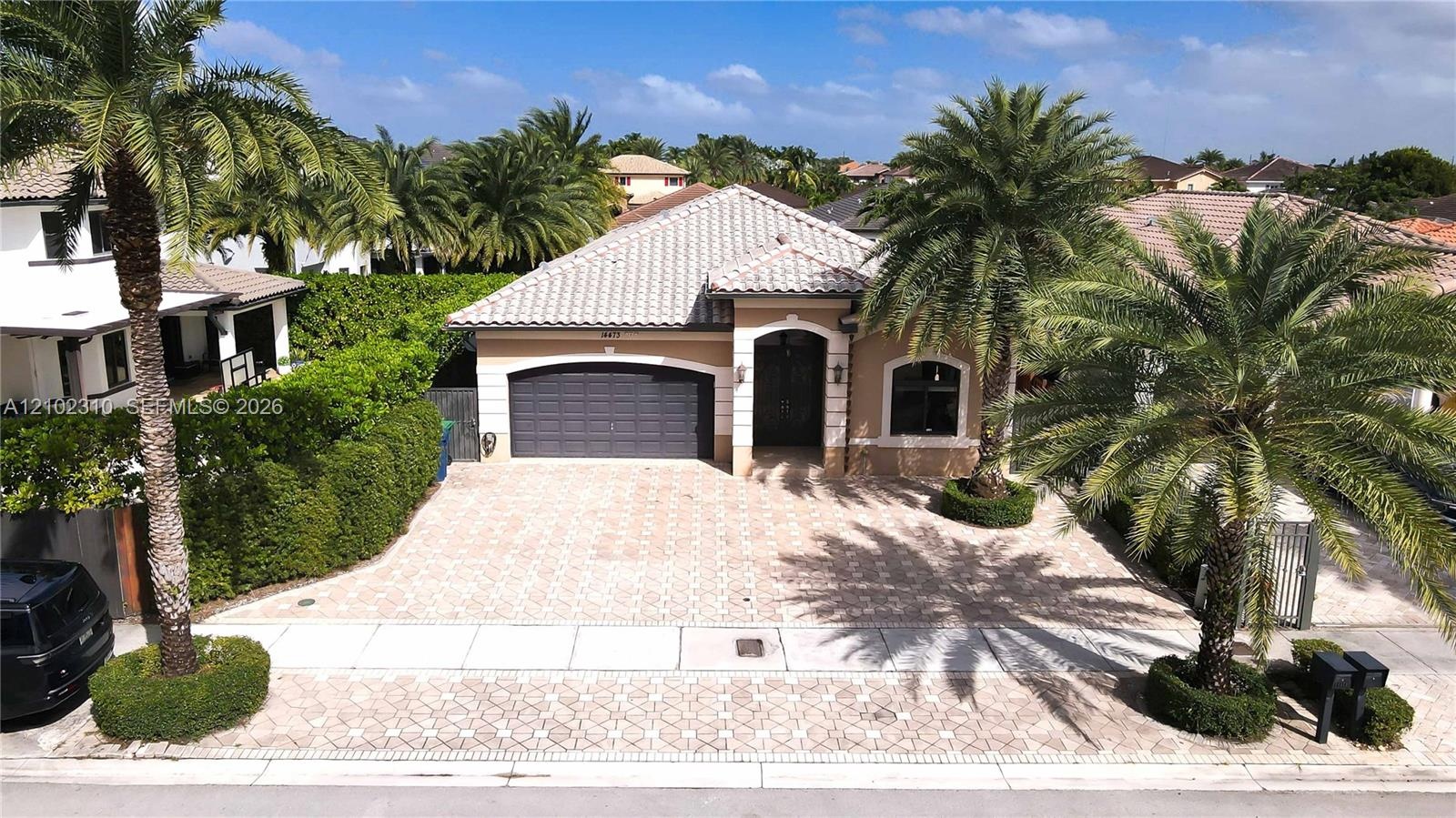 Single-story exterior featuring a concrete tile roof, paver driveway, two-car garage, and mature palm trees