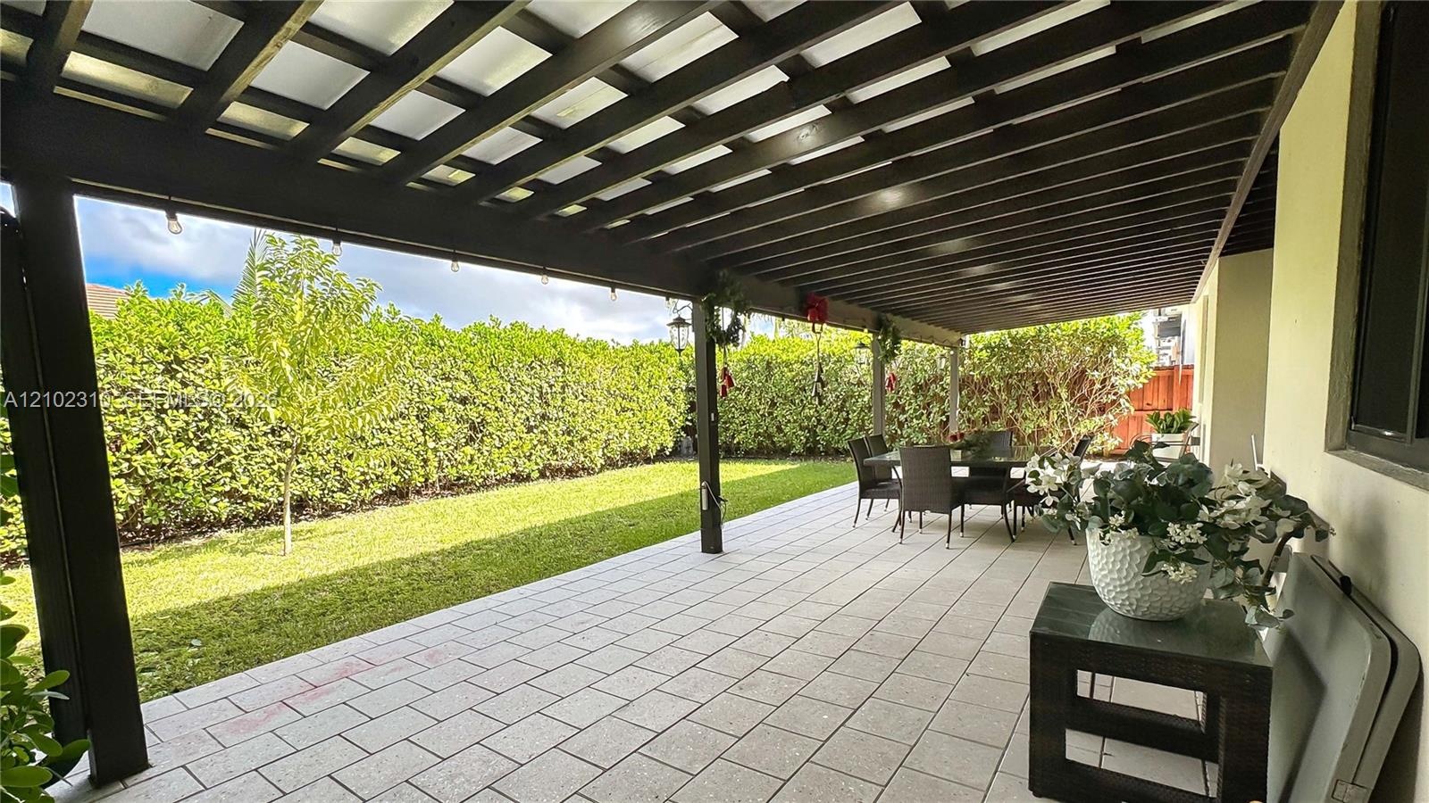 Covered patio featuring a timber-frame pergola, paver flooring, and a manicured lawn enclosed by mature privacy hedges